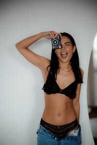 Na zdjęciu uśmiechnięta kobieta w czarnym bieliźnianym komplecie dopasowanym do sylwetki. Modelka pozuje, trzymając mały aparat fotograficzny przed twarzą.
